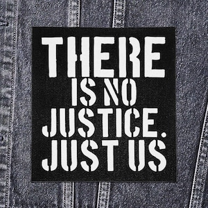 Op de afbeelding: Zwarte vierkante patch met witte tekst: "THERE IS NO JUSTICE. JUST US." De patch is op een spijkerjasje. De tekst is vetgedrukt en zonder schreef, wat een sterke boodschap overbrengt.