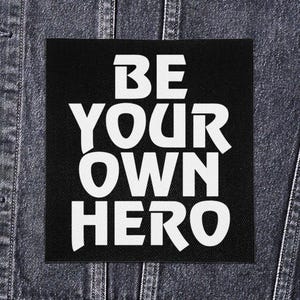 May include: A black square patch with the white text "BE YOUR OWN HERO" in a bold, sans-serif font. The patch is set against a denim jacket background, highlighting the patch's potential use as an accessory.