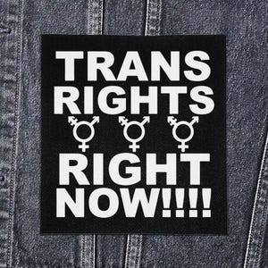Op de afbeelding: Zwart vierkantige patch met witte tekst "TRANS RIGHTS RIGHT NOW!!!!" en drie gender symbolen. De patch is op een spijkerjasje.