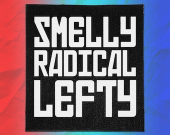 Op de afbeelding: Een vierkante zwarte stoffen patch met de witte tekst "SMELLY RADICAL LEFTY" in een vet, blokkerig schreefloos lettertype. De patch heeft een zichtbare geweven textuur en is gecentreerd tegen een achtergrond met een rood en blauw verloop.