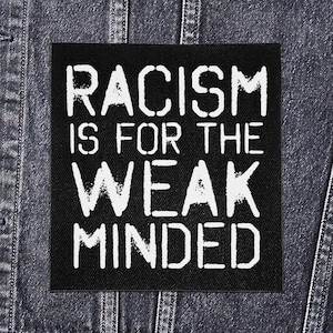 Puede incluir: Un parche cuadrado negro con el texto blanco "RACISM IS FOR THE WEAK MINDED" se muestra contra una chaqueta vaquera. El texto está en negrita, fuente sans-serif.