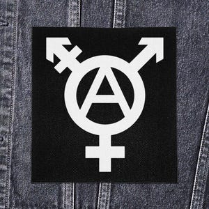 Op de afbeelding: Een zwarte vierkante patch met een wit symbool dat het transgender symbool, het anarchie symbool en het vrouwelijke symbool combineert. De patch is waarschijnlijk van stof en wordt getoond op een spijkerjasje.