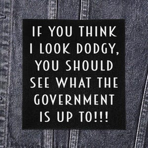 Op de afbeelding: Een zwart vierkant embleem met witte tekst: "IF YOU THINK I LOOK DODGY, YOU SHOULD SEE WHAT THE GOVERNMENT IS UP TO!!!" Het embleem is bevestigd op een spijkerjasje.