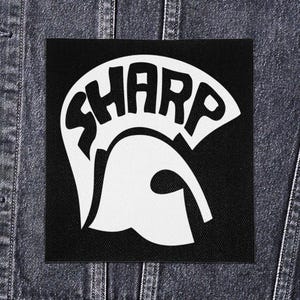 Op de afbeelding: Een zwart vierkant embleem met het woord "SHARP" in witte letters, boven een wit silhouet van een skinhead-hoofd. Het embleem is geplaatst op een spijkerjasje.