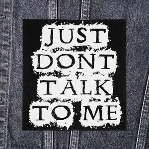 Może przedstawiać: Czarna kwadratowa naszywka z białym napisem "JUST DONT TALK TO ME" w stylu distressed. Naszywka jest umieszczona na dżinsowej kurtce.