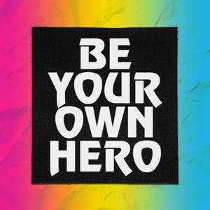 May include: A square black fabric patch featuring the white text BE YOUR OWN HERO in a bold, sans-serif font. The patch has a visible woven texture and is centered against a vibrant, multi-colored background with a gradient of rainbow colors.