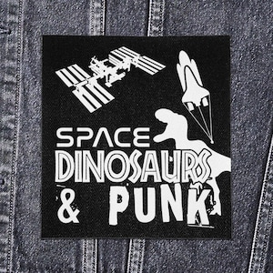 May include: Black square patch featuring a white graphic of the International Space Station, a rocket, and a dinosaur silhouette. Text reads "SPACE DINOSAURS & PUNK". The patch is set against a denim background.