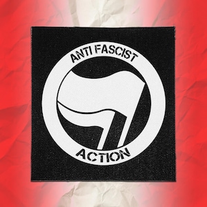 May include: A square black fabric patch featuring a white circular emblem with two waving flags inside. The text ANTI FASCIST is printed along the top curve of the circle, and the word ACTION is printed along the bottom curve in a distressed font.
