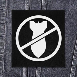 Op de afbeelding: Een zwarte vierkante patch met een witte afbeelding van een bom in een cirkel met een diagonale lijn erdoor. De patch zit op een spijkerjasje.