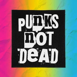 May include: A square black fabric patch featuring the white text PUNKS NOT DEAD in a bold, distressed, block-letter font. The lettering has a rough, stencil-like appearance with visible texture, set against a solid black background with clean edges.