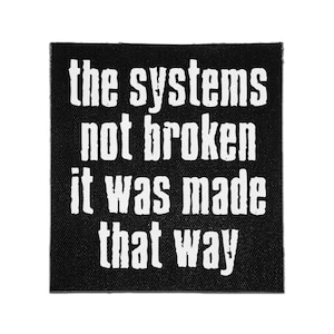 May include: A square black fabric patch featuring white distressed-style lettering that reads: the systems not broken it was made that way. The text is arranged in four lines, centered on the dark background with a visible woven textile texture.