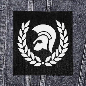 Op de afbeelding: Zwart vierkantige patch met een wit Trojaans helm embleem omcirkeld door een lauwerkrans. De patch is geplaatst tegen een donkere denim achtergrond, met een klassiek ontwerp.