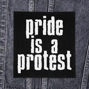 以下が含まれることがあります： 「pride is a protest」の白文字がプリントされた黒い正方形のパッチ。デニムジャケットに表示されています。テキストは太字のサンセリフ体です。