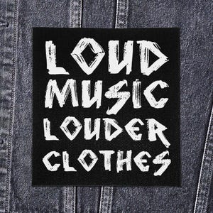 May include: Black square patch with white text that reads "LOUD MUSIC LOUDER CLOTHES." The patch is affixed to a denim jacket. The text is in a bold, distressed font, suggesting a punk or rock aesthetic.