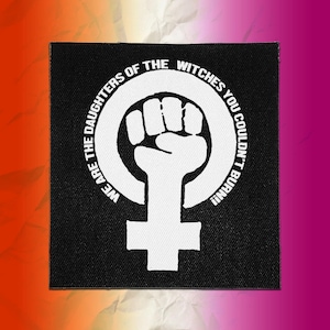May include: A square black fabric patch featuring a white graphic of a raised fist inside a female gender symbol. The text "WE ARE THE DAUGHTERS OF THE WITCHES YOU COULDN'T BURN!!" is printed in white capital letters along the circular border.