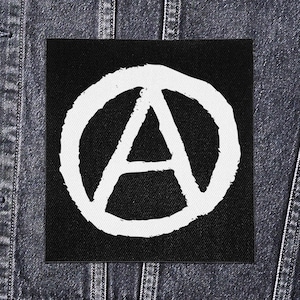 May include: A black square patch featuring a white anarchy symbol, an "A" inside a circle. The patch is set against a dark denim background, with visible stitching and seams.