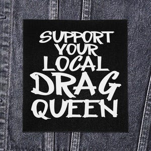 Puede incluir: Un parche cuadrado negro con texto blanco que dice "SUPPORT YOUR LOCAL DRAG QUEEN". El parche se muestra en una chaqueta vaquera. El texto está en un estilo de escritura a mano en negrita.