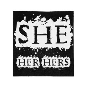May include: A square black fabric patch featuring a white distressed paint-style rectangular background. The word SHE is printed in large black serif letters, with the words HER and HERS printed below in smaller black serif letters.