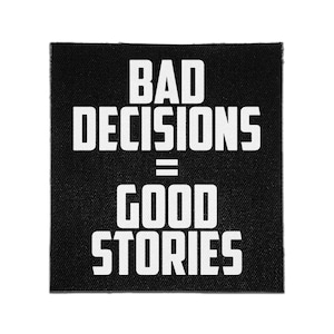 May include: A square black fabric patch featuring the white text BAD DECISIONS = GOOD STORIES in a bold, sans-serif font. The design is centered on the patch, which has a visible woven texture throughout the black background and white lettering.