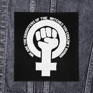 May include: Black square patch with a white graphic of a raised fist inside a circle, with the female symbol at the bottom. The text "WE ARE THE DAUGHTERS OF THE WITCHES YOU COULDN'T BURN!!" is printed around the circle.