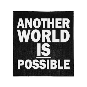 May include: A square black fabric patch featuring the white text "ANOTHER WORLD IS POSSIBLE" in a bold, sans-serif font. The design includes a horizontal white line positioned between the words "IS" and "POSSIBLE" on the black background.