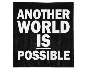 May include: A square black fabric patch featuring the white text "ANOTHER WORLD IS POSSIBLE" in a bold, sans-serif font. The design includes a horizontal white line positioned between the words "IS" and "POSSIBLE" on the black background.