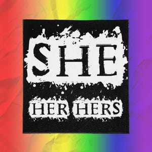 May include: A square black fabric patch featuring a white distressed paint-style rectangular background. The word SHE is printed in large black serif letters, with the words HER HERS printed below in smaller black serif letters on a white base.
