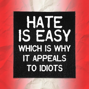 May include: A square black fabric patch featuring white embroidered text that reads HATE IS EASY WHICH IS WHY IT APPEALS TO IDIOTS. The lettering is in a bold, slightly distressed font style, centered on the dark background of the patch.
