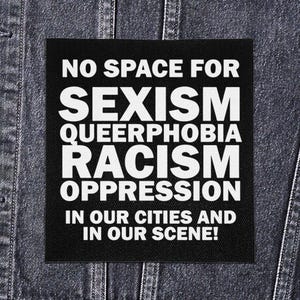 May include: A black square patch with white text. The text reads "NO SPACE FOR SEXISM QUEERPHOBIA RACISM OPPRESSION IN OUR CITIES AND IN OUR SCENE!" The patch is set against a denim background.