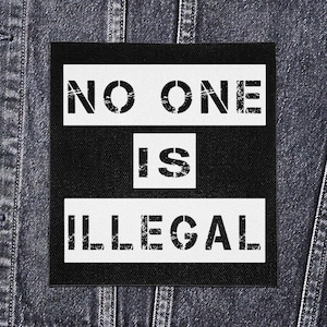 Puede incluir: Un parche cuadrado con las palabras "NO ONE IS ILLEGAL" en letras blancas, estilo plantilla, sobre un fondo negro. El parche está colocado sobre una chaqueta vaquera.
