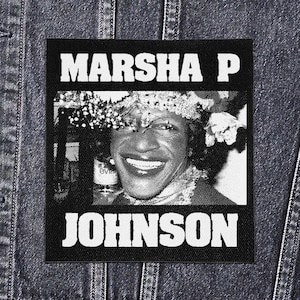 Pode incluir: Patch em preto e branco com o texto "MARSHA P" acima de uma imagem de uma pessoa sorrindo com uma coroa de flores e "JOHNSON" abaixo. O texto está em letras brancas em negrito.