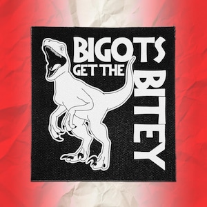 May include: A square black fabric patch featuring a white line art illustration of a dinosaur with its mouth open. The text BIGOTS GET THE BITEY is printed in white capital letters next to the dinosaur figure on the black background.