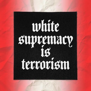 Może przedstawiać: Kwadratowa czarna naszywka z tkaniny z białym napisem w stylu gotyckim o treści "white supremacy is terrorism" wyśrodkowanym na powierzchni. Naszywka ma widoczną tkaną fakturę i proste krawędzie, co zapewnia wyraźny kontrastowy wygląd.