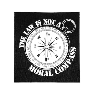 Può includere: Una toppa quadrata in tessuto nero con una grafica bianca di una bussola al centro. Il testo "THE LAW IS NOT A MORAL COMPASS" è stampato in lettere maiuscole bianche, curvate attorno alla parte superiore e inferiore dell'illustrazione.