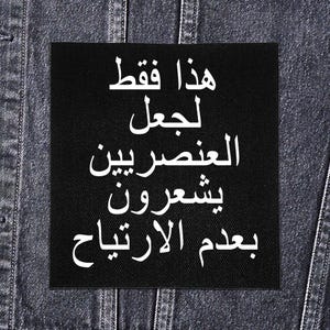 Op de afbeelding: Een zwart vierkant patch met witte Arabische tekst, genaaid op een donker spijkerjasje. De tekst luidt: "Dit is alleen om racisten ongemakkelijk te maken."
