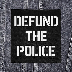 May include: Black square patch with the words "DEFUND THE POLICE" in bold white stencil font. The patch is set against a denim jacket background.