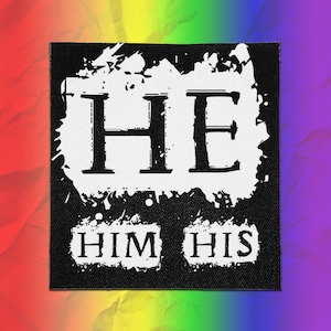 May include: A square black fabric patch featuring a white ink splatter design. The text HE is printed in large black serif letters within the top splatter, while the words HIM and HIS appear in smaller black serif letters below.