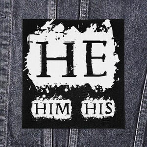Op de afbeelding: Zwart-witte vierkante patch met de woorden "HE" in grote hoofdletters en "HIM HIS" eronder. De tekst is in een vetgedrukte, schreefloze lettertype. De patch is geplaatst op een spijkerjasje.