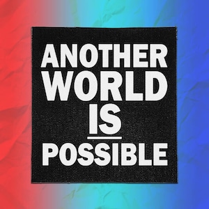 May include: A square black fabric patch featuring the white text "ANOTHER WORLD IS POSSIBLE" in a bold, sans-serif font. The text is arranged in four lines with a horizontal line separating the words "IS" and "POSSIBLE" on the patch surface.