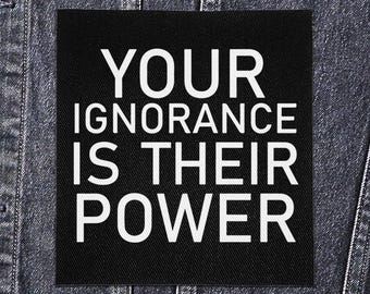 May include: Black square patch with the white text "YOUR IGNORANCE IS THEIR POWER" sewn onto a denim jacket. The text is in a bold, sans-serif font.