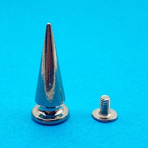 May include: A silver-coloured metal cone-shaped stud with a smooth, reflective finish. The stud features a pointed top and a circular base. Beside the stud is a separate, smaller silver-coloured metal screw component with a threaded post and flat base.