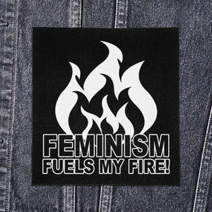 Puede incluir: Parche cuadrado negro con un diseño de llama blanca y el texto "FEMINISM FUELS MY FIRE!". El parche es probablemente de tela y se muestra en una chaqueta vaquera.