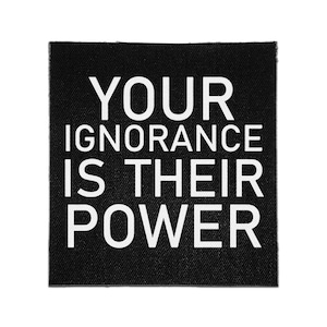 May include: A square black fabric patch featuring the white text YOUR IGNORANCE IS THEIR POWER printed in a bold, sans-serif font. The patch has a visible woven texture and clean, straight edges, designed for attachment to clothing or accessories.