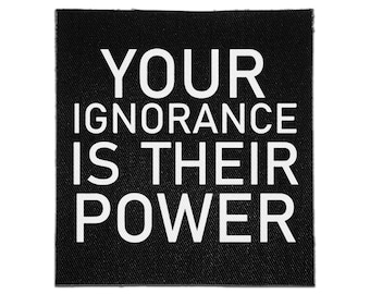 May include: A square black fabric patch featuring the white text YOUR IGNORANCE IS THEIR POWER printed in a bold, sans-serif font. The patch has a visible woven texture and clean, straight edges, designed for attachment to clothing or accessories.
