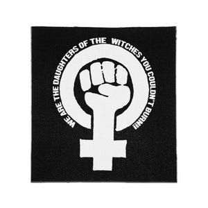 May include: A square black fabric patch featuring a white screen-printed design of a raised fist inside a female gender symbol. The text "WE ARE THE DAUGHTERS OF THE WITCHES YOU COULDN'T BURN!!" is printed in a circular arc around the central graphic.