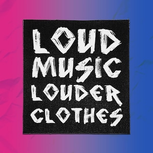 Pode incluir: Um emblema quadrado de tecido preto com o texto branco LOUD MUSIC LOUDER CLOTHES numa fonte desgastada de traço irregular. As letras estão dispostas em quatro linhas horizontais centradas no fundo preto com uma textura tecida visível.