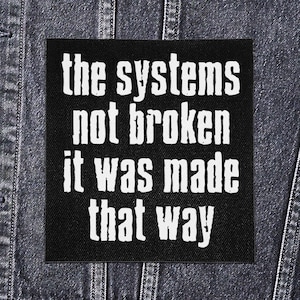 Puede incluir: Parche cuadrado negro con texto blanco que dice "the systems not broken it was made that way". El parche está sobre una chaqueta vaquera.