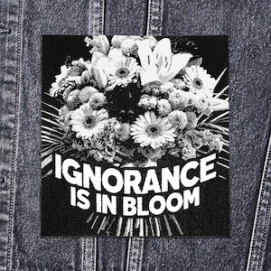 May include: A black and white patch with a floral bouquet design. The text "IGNORANCE IS IN BLOOM" is displayed in a bold font. The patch is square and made of fabric. The image is set against a denim background.