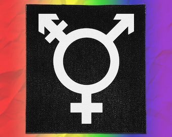 May include: A square black fabric patch featuring a white embroidered transgender symbol centered on the surface. The symbol consists of a circle with a cross, an arrow, and a combined cross and arrow pointing in different directions.