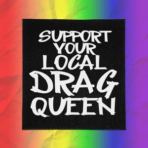 May include: A square black fabric patch featuring the white text SUPPORT YOUR LOCAL DRAG QUEEN in a bold, stylized font. The patch has a visible woven texture and is centered against a vibrant, multi-colored rainbow gradient background.